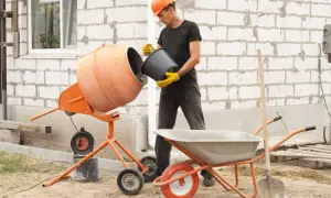 Budowlaniec w kasku wsypuje materiał do betoniarki. Obok stoi taczka i łopata.
