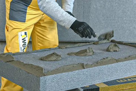 Nakładanie kleju do styropianu grafitowego na płytę izolacyjną. Ręce w czarnych rękawiczkach rozprowadzają masę szpachlą.